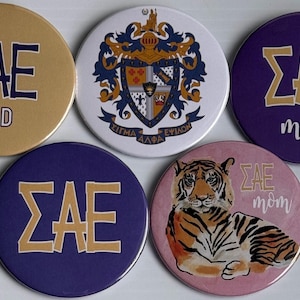 May include: Five circular buttons featuring Sigma Alpha Epsilon fraternity designs. One is gold with blue text, one is white with a crest, two are purple with gold text, and one is pink with a tiger illustration and the text SAE mom.