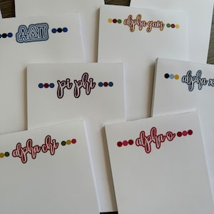 May include: A collection of white notepads featuring various sorority and fraternity names in colorful cursive fonts. Each notepad has a row of colored dots above or below the text, with a wooden surface in the background.