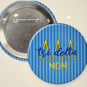 Puede incluir: Botón a rayas azules y amarillas con tres triángulos amarillos y el texto "tri delta mom" en azul.