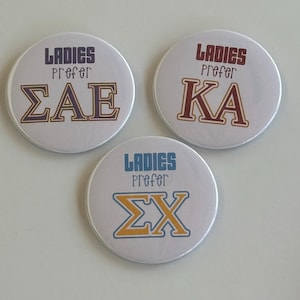 Op de afbeelding: Drie ronde witte knoppen met de tekst "Ladies Prefer" en een symbool voor een studentenvereniging. De symbolen zijn "SAE", "KA" en "ΣX".
