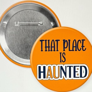 Könnte beinhalten: Orangener Knopf mit dem Text "THAT PLACE IS HAUNTED" in schwarzer und blauer Schrift. Die Rückseite des Knopfes ist silber.