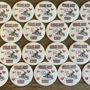 May include: A collection of 16 round white buttons with black and pink text and graphics. Each button features a different Greek letter sorority name and the text "Game Day the ... Way".