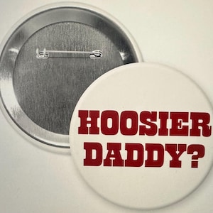 May include: A white button with the text "HOOSIER DADDY?" in red, bold letters. The button is round and has a pin back. Another button is visible in the background.