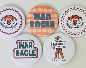 May include: A collection of five round buttons with Auburn University designs. The buttons feature the words "WAR EAGLE", a tiger mascot, and the phrase "Let's Go!" in orange, navy, and white.