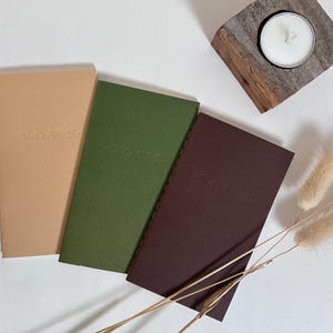 May include: Three notebooks in tan, green, and burgundy, with the word "VOWS" embossed on the tan notebook. A small candle sits in a wooden holder. Dried flowers are arranged near the notebooks.