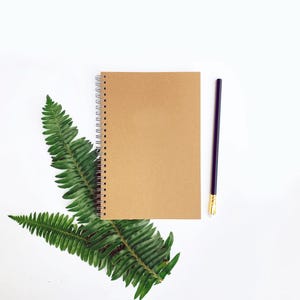 May include: A spiral-bound notebook with a kraft paper cover rests on a bed of green fern leaves. A black pencil with a gold-colored ferrule lies to the right. The items are arranged on a white background, creating a minimalist, nature-inspired aesthetic.
