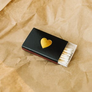 May include: A black matchbox with a gold heart design. The matchbox is open and contains white tipped matches.