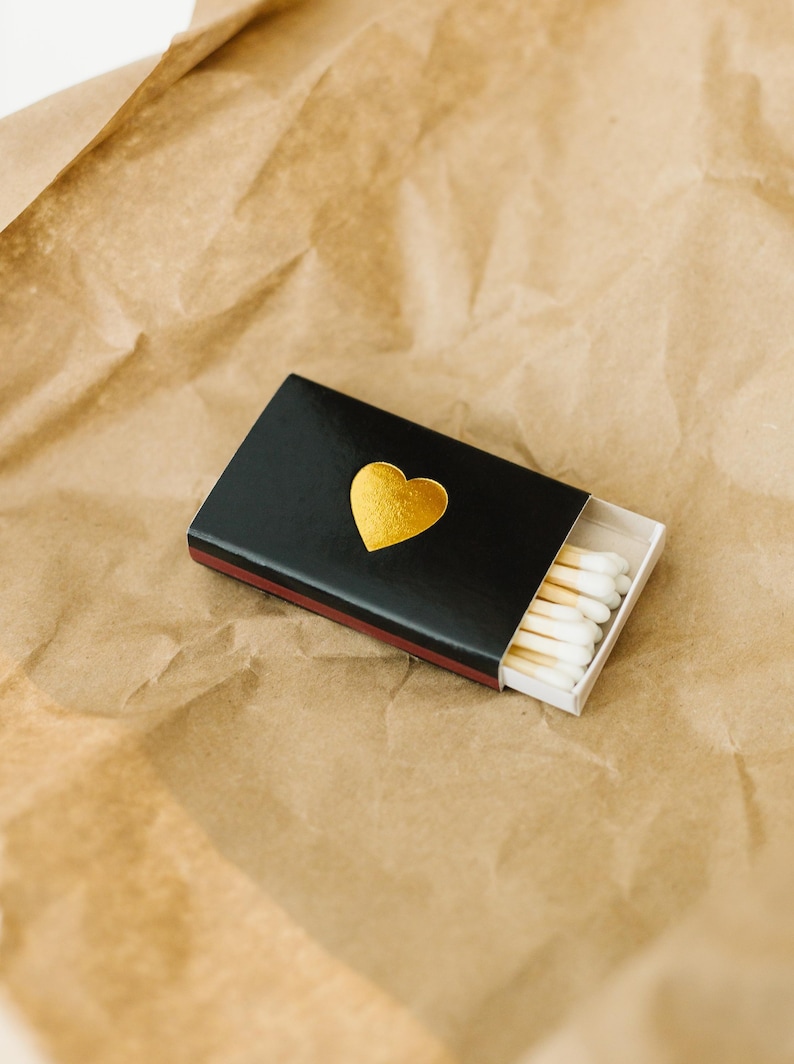 May include: A black matchbox with a gold heart design. The matchbox is open and contains white tipped matches.