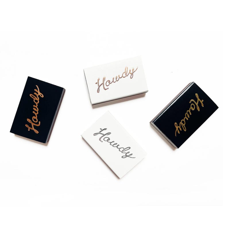 May include: Four matchboxes with the word "Howdy" written on them in gold script. Two matchboxes are black with white lettering and two are white with gold lettering.