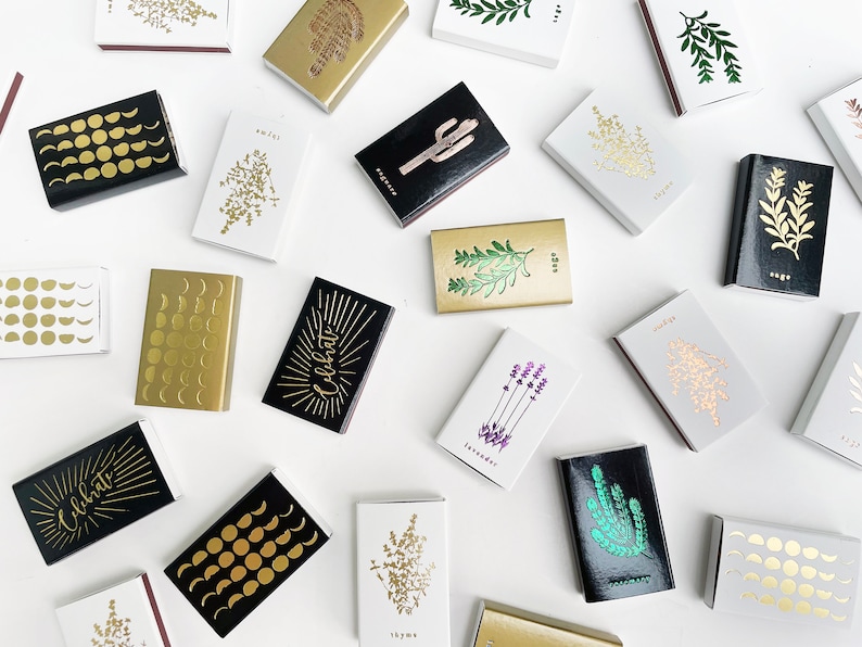 May include: A collection of matchboxes with various designs. The matchboxes feature gold and black patterns, including moons, stars, and botanical illustrations. Some matchboxes have the word "Celebrate" printed on them.