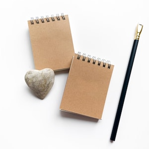 Puede incluir: Dos pequeños cuadernos con espiral y cubiertas de papel kraft se colocan cerca de una piedra en forma de corazón y un lápiz negro con una virola dorada. Los artículos están dispuestos sobre una superficie blanca.