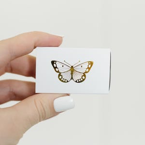 May include: A white box with a gold butterfly design. The butterfly is centered on the box and has a detailed design.