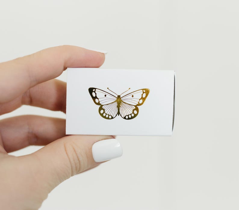 May include: A white box with a gold butterfly design. The butterfly is centered on the box and has a detailed design.