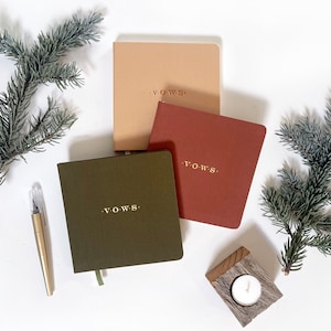 May include: Three square notebooks in tan, terracotta, and olive green, each featuring the word V.O.W.S. embossed in gold on the front cover. The set includes a gold pen, two evergreen branches, and a small tea light candle in a wooden holder.
