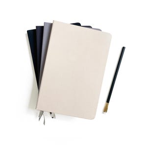 Pode incluir: Uma pilha de cinco cadernos de capa dura em várias cores: creme, cinza, azul marinho e preto. Cada caderno tem uma fita de marcador. Um lápis preto com ponta dourada repousa ao lado dos cadernos sobre um fundo branco.
