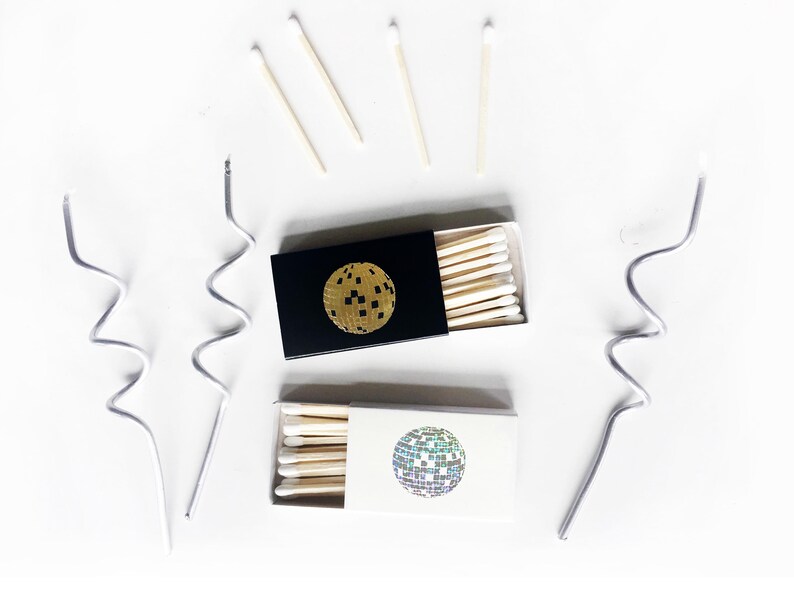 May include: Two boxes of matches with a disco ball design. One box is black with a gold disco ball and the other is white with a silver disco ball.