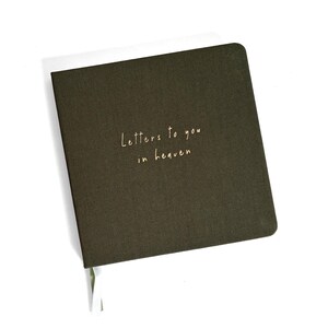 May include: A square, dark olive green journal with a fabric-textured cover. The front cover features the gold-colored text "Letters to you in heaven" printed in a simple, handwritten-style font. A small white ribbon bookmark extends from the bottom.