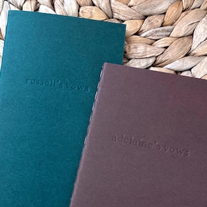 Op de afbeelding: Twee rechthoekige gelofteboekjes met een matte kaft. Het langere boekje is donkergroen met de in reliëf gedrukte tekst "russell's vows" in het midden. Het kortere boekje is donkerbruin met de in reliëf gedrukte tekst "adelaine's vows" in het midden.