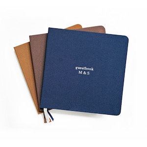 May include: Three hardcover guestbooks in shades of blue, brown, and tan are fanned out on a white background. The navy blue book on top has the word "guestbook" and the initials "M & S" printed on the cover. A ribbon bookmark is attached.