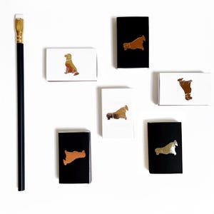 May include: Six small matchboxes with different dog breeds printed on them. The matchboxes are white, black, and brown with gold foil accents. The matchboxes are arranged in a row on a white surface.