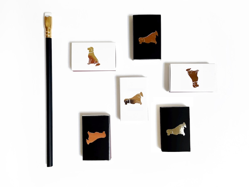 May include: Six small matchboxes with different dog breeds printed on them. The matchboxes are white, black, and brown with gold foil accents. The matchboxes are arranged in a row on a white surface.