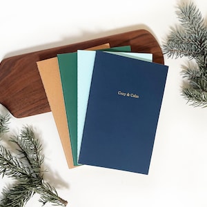 May include: A stack of notebooks in shades of navy, green, light blue, and tan. The navy notebook has the gold text "Cozy & Calm". The notebooks are arranged on a wooden cutting board with a sprig of evergreen on the side.