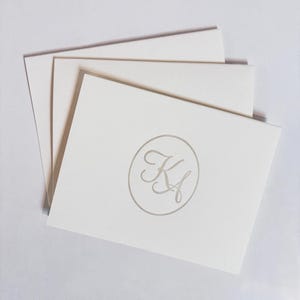 Puede incluir: Un conjunto de tres tarjetas de notas rectangulares blancas, cada una con un emblema circular gris claro centrado que contiene las letras del monograma KA en cursiva estilizada, dispuestas en una orientación vertical superpuesta.