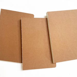 Puede incluir: Tres cuadernos marrones de diferentes tamaños están dispuestos sobre una superficie blanca. Los cuadernos tienen un diseño sencillo y minimalista con una cubierta lisa y texturizada. La encuadernación es visible en un cuaderno.