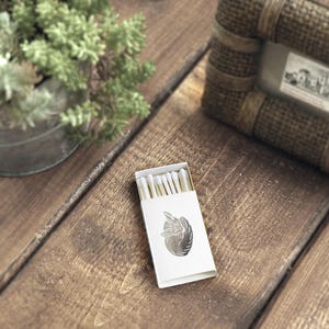 May include: A white box of matches with a silver design of a hand holding a heart on the front. The matches are visible through a clear plastic window on the box.