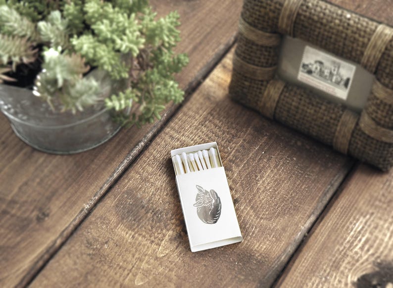 May include: A white box of matches with a silver design of a hand holding a heart on the front. The matches are visible through a clear plastic window on the box.