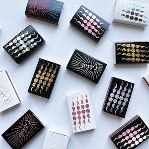 May include: A collection of matchboxes with various designs. Some boxes feature a moon phase pattern with gold, pink, or iridescent glitter. Others have the word "Celebrate" in a black and white design with a sunburst pattern.