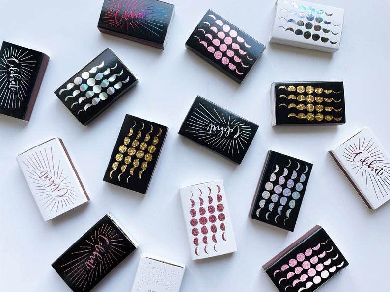 May include: A collection of matchboxes with various designs. Some boxes feature a moon phase pattern with gold, pink, or iridescent glitter. Others have the word "Celebrate" in a black and white design with a sunburst pattern.