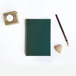 May include: A dark green spiral-bound notebook sits on a white surface, accompanied by a wooden candle holder with a lit candle, a dark pencil, and a heart-shaped stone. The notebook has a solid, deep green cover.