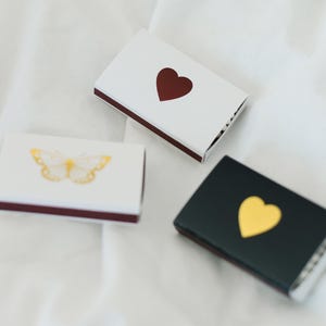 May include: Three matchboxes with different designs. One has a white background with a red heart, another has a black background with a gold heart, and the last one has a white background with a gold butterfly.