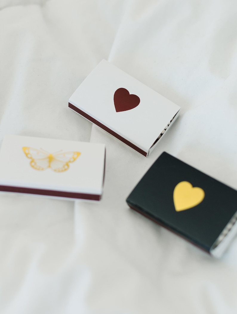 May include: Three matchboxes with different designs. One has a white background with a red heart, another has a black background with a gold heart, and the last one has a white background with a gold butterfly.