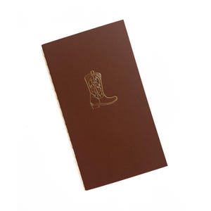 May include: A brown card with a gold-colored illustration of a cowboy boot. The boot design is detailed with decorative stitching. The card is rectangular and has a matte finish.
