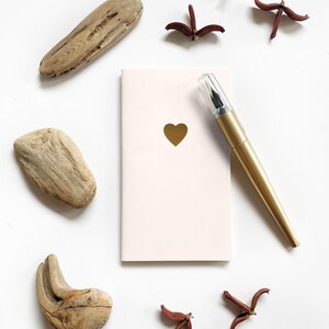 Op de afbeelding: Een zachtroze notitieboek met een gouden hart, samen met een gouden pen met een transparante dop. De compositie omvat stukken drijfhout en gedroogde bloemelementen, alles op een witte achtergrond.