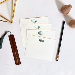 Pode incluir: Um conjunto de papelaria com quatro cartões de notas brancos com um monograma verde-azulado, um marcador de couro castanho com uma borla verde, um recipiente geométrico dourado, duas peças decorativas em forma de concha de madeira e um lápis verde.