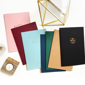 May include: A set of six notebooks with different coloured covers. The covers are pink, burgundy, light blue, navy blue, green, and black. The notebooks have gold lettering and a gold logo on the covers. The notebooks are arranged in a fan shape on a white surface.