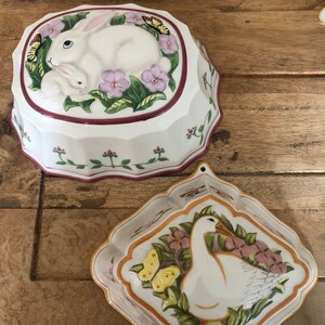 Puede incluir: Dos moldes de cerámica para hornear postres. Un molde presenta un conejo blanco con un conejito y flores rosas. El otro molde presenta un ganso blanco con mariposas amarillas y flores rosas.
