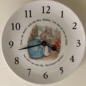 A white ceramic circular clock in the style of a plate. Black numerals and hands with central image an illustration by Beatrix Potter of Mrs Rabbit doing up Peter Rabbits blue jacket and the three Flopsy bunnies in the background in their red capes.