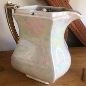 以下が含まれることがあります： 金色のハンドルとトリムが付いた、虹色の白いセラミックピッチャー。ピッチャーはユニークな角張った形状で、注ぎ口が付いています。ハンドルは湾曲した金色です。ピッチャーはおそらくヴィンテージで、テーブルセッティングにエレガンスを加えます。
