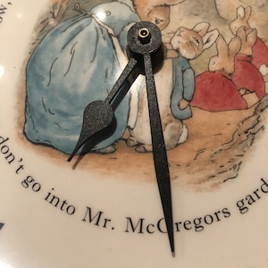 A close up of the black clock hands and the image of Mrs Rabbit fastening Peter Rabbits blue jacket. The three Flopsy bunnies in their red capes are in the background on their way towards the woods. Mrs Rabbit wears a blue dress and white apron