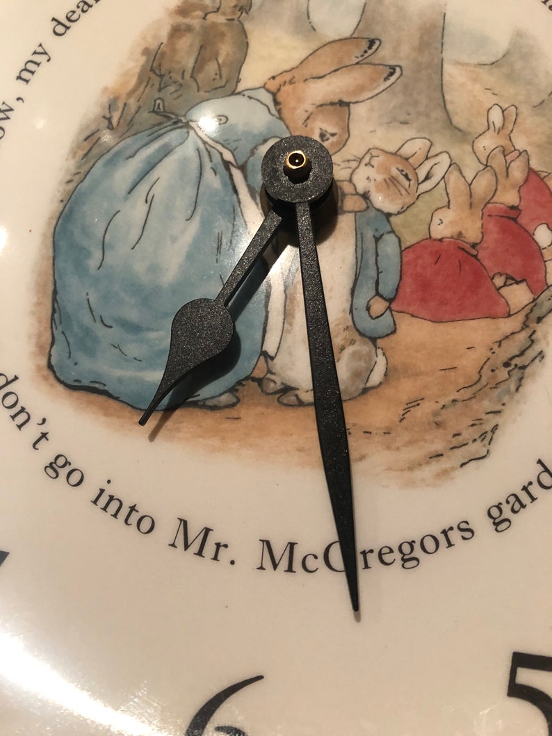 A close up of the black clock hands and the image of Mrs Rabbit fastening Peter Rabbits blue jacket. The three Flopsy bunnies in their red capes are in the background on their way towards the woods. Mrs Rabbit wears a blue dress and white apron