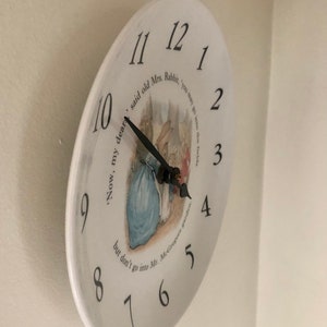 A side view of the white circular ceramic plate showing the numerals around the face the black clock hands and the central illustration of Mrs Rabbit Peter Rabbit and the Flopy Bunnies