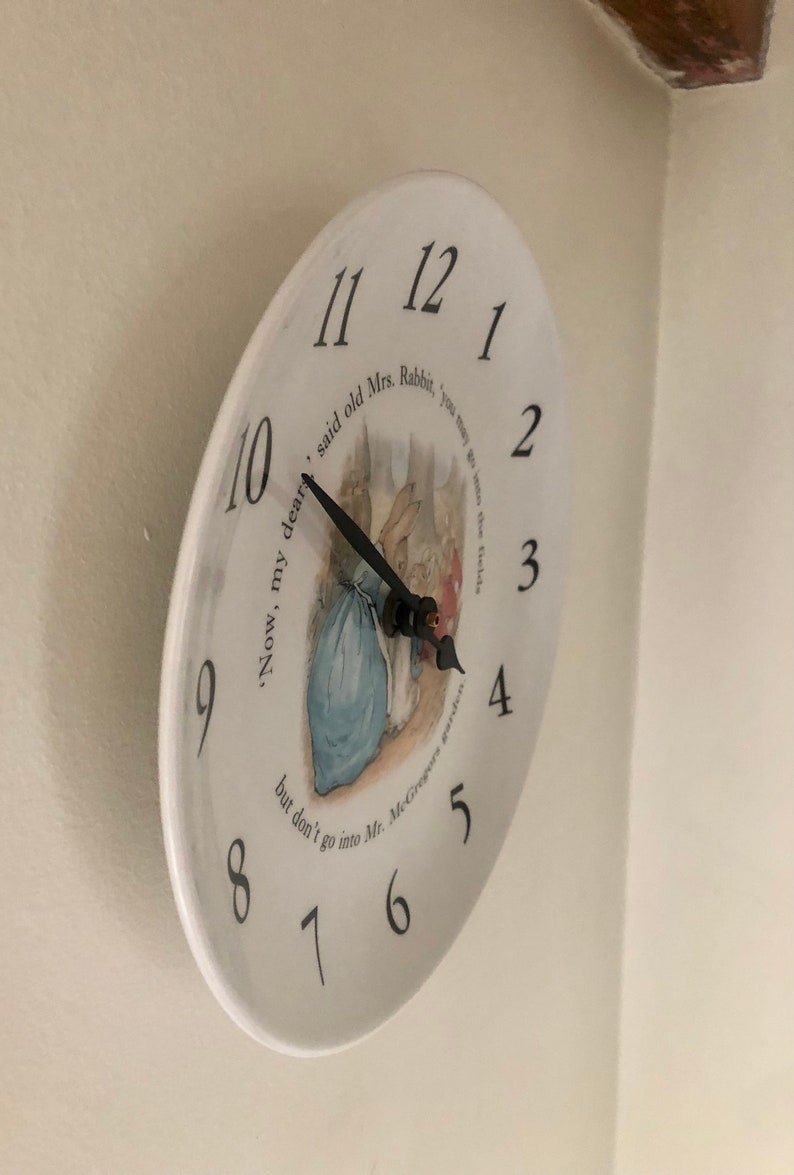 A side view of the white circular ceramic plate showing the numerals around the face the black clock hands and the central illustration of Mrs Rabbit Peter Rabbit and the Flopy Bunnies