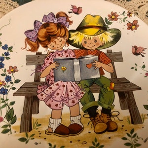 Può includere: Un piatto in ceramica vintage con un'illustrazione colorata di due bambini seduti su una panchina di legno che leggono un libro insieme. Il piatto ha uno sfondo bianco con motivi floreali e il testo "Gott schlich" in basso.