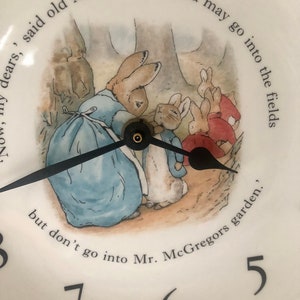 A close up of the black clock hands and the image of Mrs Rabbit fastening Peter Rabbits blue jacket. The three Flopsy bunnies in their red capes are in the background on their way towards the woods. A phrase from the book can be seen around the image