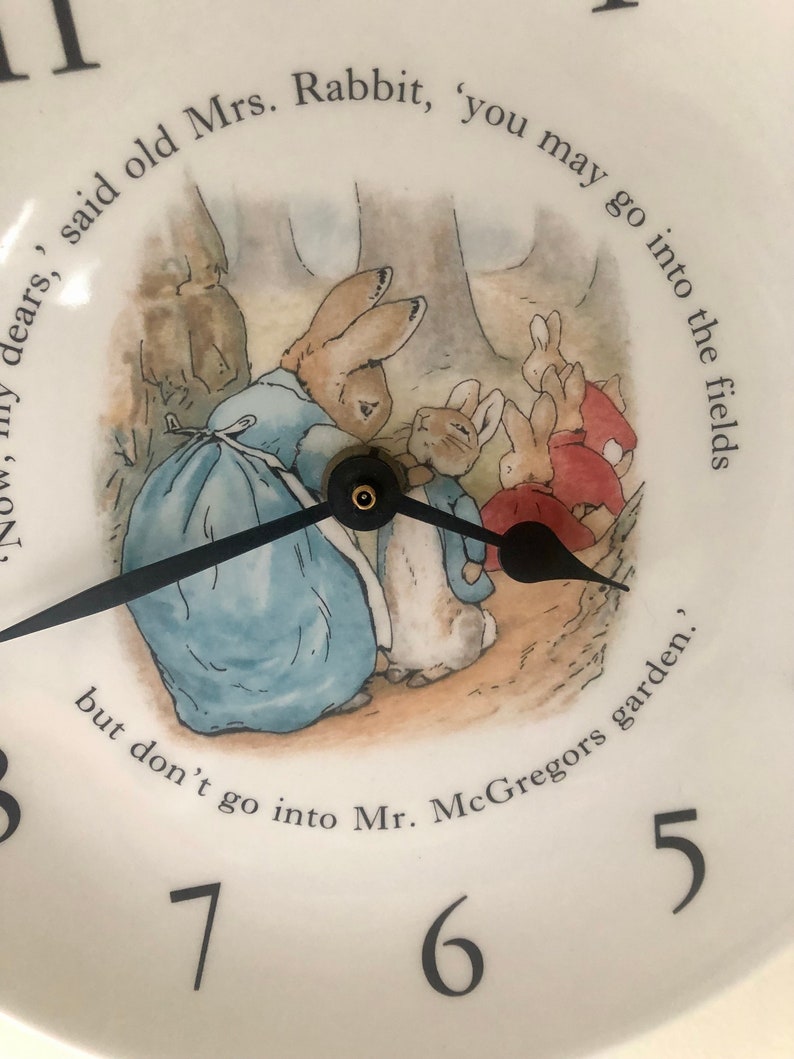 A close up of the black clock hands and the image of Mrs Rabbit fastening Peter Rabbits blue jacket. The three Flopsy bunnies in their red capes are in the background on their way towards the woods. A phrase from the book can be seen around the image