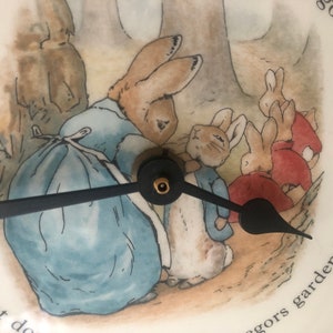 A close up of the black clock hands and the image of Mrs Rabbit fastening Peter Rabbits blue jacket. The three Flopsy bunnies in their red capes are in the background on their way towards the woods. Mrs Rabbit wears a blue dress and white apron